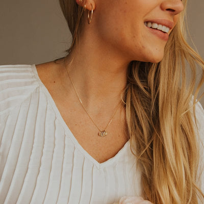 A close-up of a blonde woman in a white long sleeve pleated top wearing a delicate gold pendant chain with four round flat engraved disc charms, set against a dark grey background.