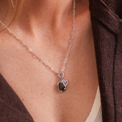 A close-up of a person wearing a silver paperclip link chain necklace with a round ring in the center covered in clear stones, holding a silver oval locket and a silver heart charm.