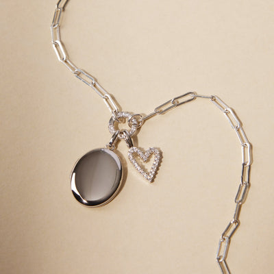 A silver paperclip link chain necklace with a round ring in the center covered in clear stones, holding a silver oval locket charm and a silver heart charm embellished with clear gemstones.