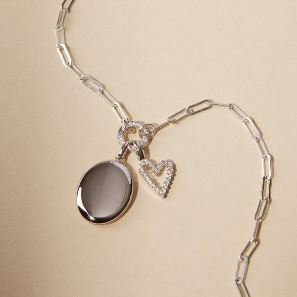 A silver paperclip link chain necklace with a round ring in the center covered in clear stones, holding a silver oval locket charm and a silver heart charm embellished with clear gemstones.