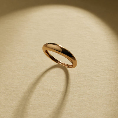A polished gold band ring with a smooth, rounded organic shape, displayed on a textured beige background with natural shadows.