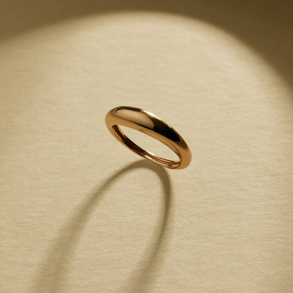 A polished gold band ring with a smooth, rounded organic shape, displayed on a textured beige background with natural shadows.