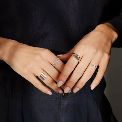 A close-up of a person's hands wearing multiple silver rings, including multiple stacked rings with clear gemstones, a wide cigar band ring, a thin polished ring, and two silver bubble rings.