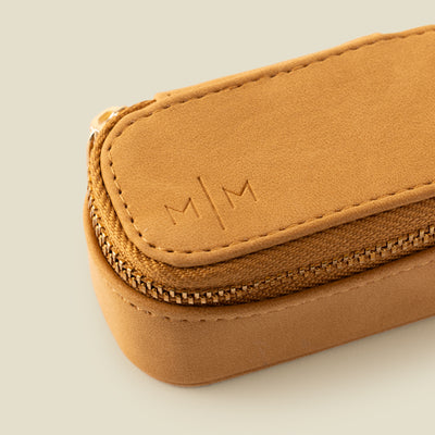A close-up of a  small tan leather zippered jewelry case with an embossed 'M|M' logo on the top, photographed on a solid light beige background.