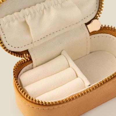 A close-up of the cream interior of a small jewelry case, showcasing three slots for rings, and open compartment, and a small pocket on the lid.