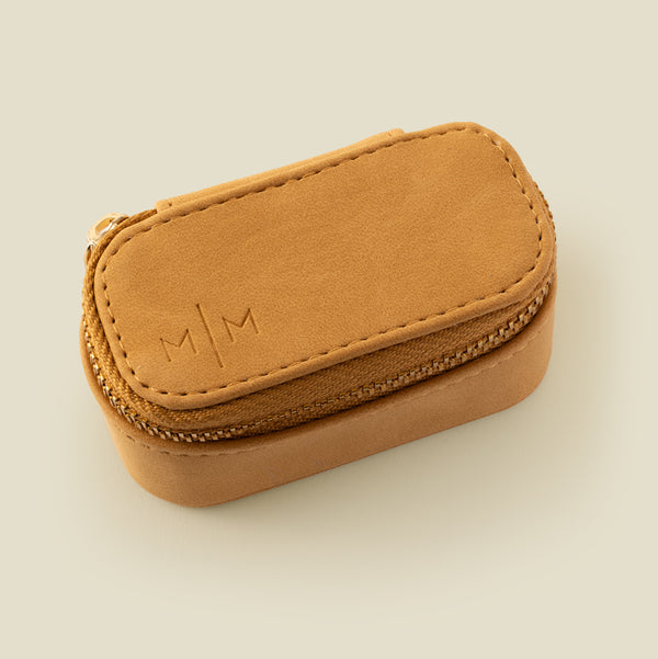 A small tan leather zippered jewelry case with an embossed 'M|M' logo on the top, photographed on a solid light beige background.