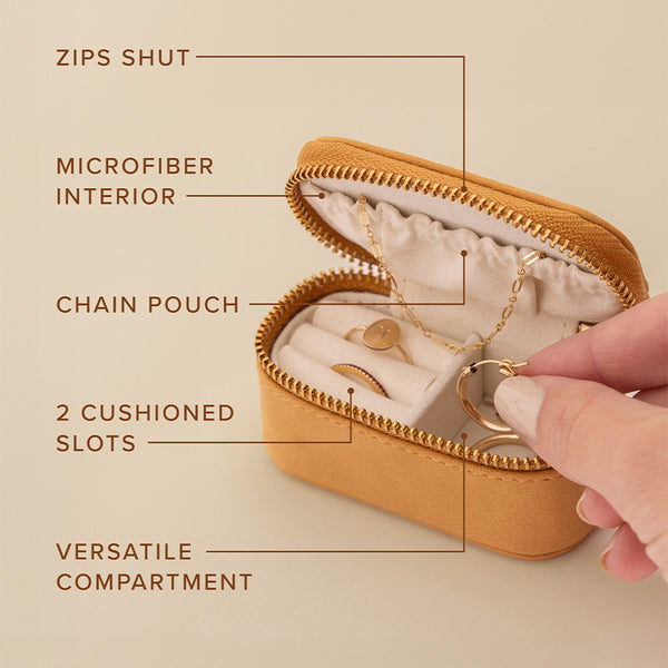 A small tan leather zippered jewelry case shown open with a hand reaching in to grab jewelry out. Over the image are multiple tag lines with lines pointing out features of the case, including 'ZIPS SHUT', 'MICROFIBER INTERIOR', 'CHAIN POUCH', '2 CUSHIONED SLOTS', and 'VERSATILE COMPARTMENT'.