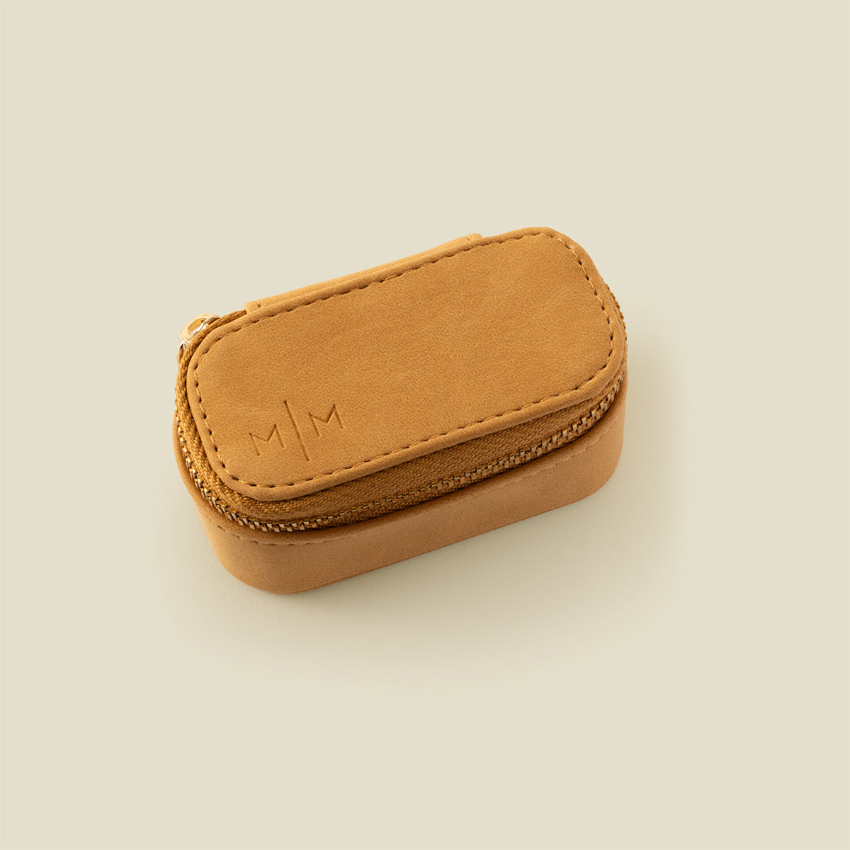 A small tan leather zippered jewelry case with an embossed 'M|M' logo on the top, photographed on a solid light beige background.