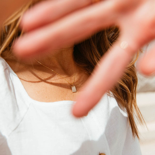 A hand half covering the camera, showing a person in a white top wearing a gold pendant necklace with a rectangle charm.