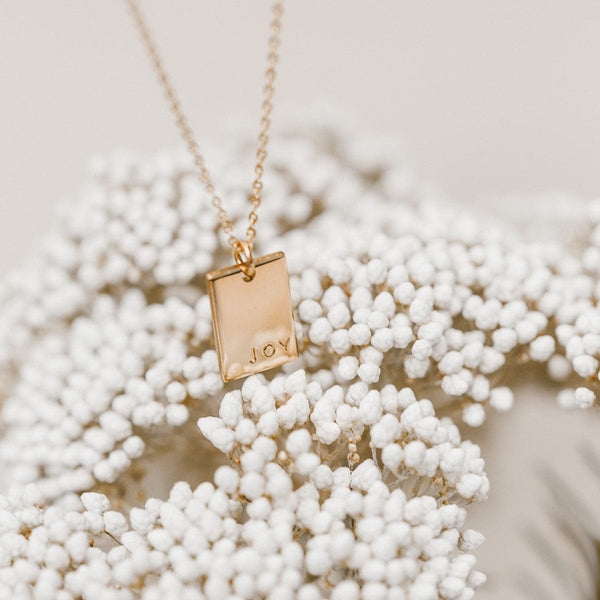 A delicate gold pendant chain necklace with a small rectangular charm engraved with the word 'JOY',  hanging on top of small white flowers with a brown stem.