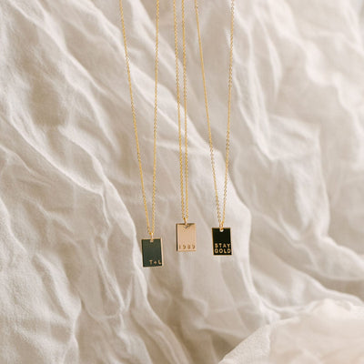 Three gold delicate pendant necklaces, each with a small flat rectangular charm with different words engraved, hanging in front of a textured white linen background.