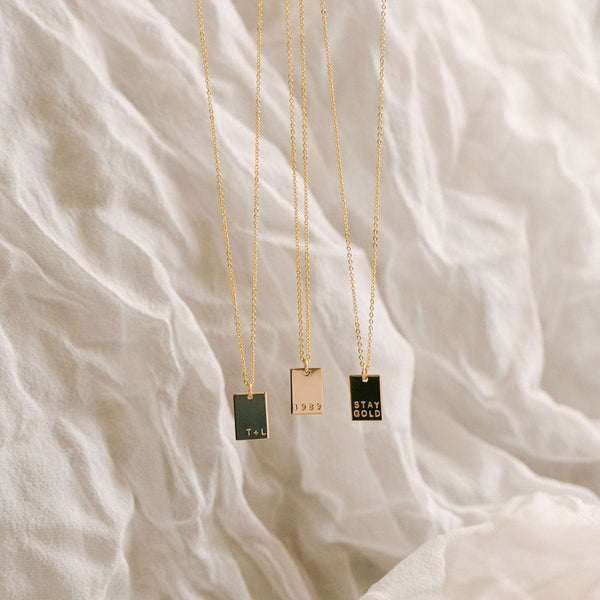 Three gold delicate pendant necklaces, each with a small flat rectangular charm with different words engraved, hanging in front of a textured white linen background.