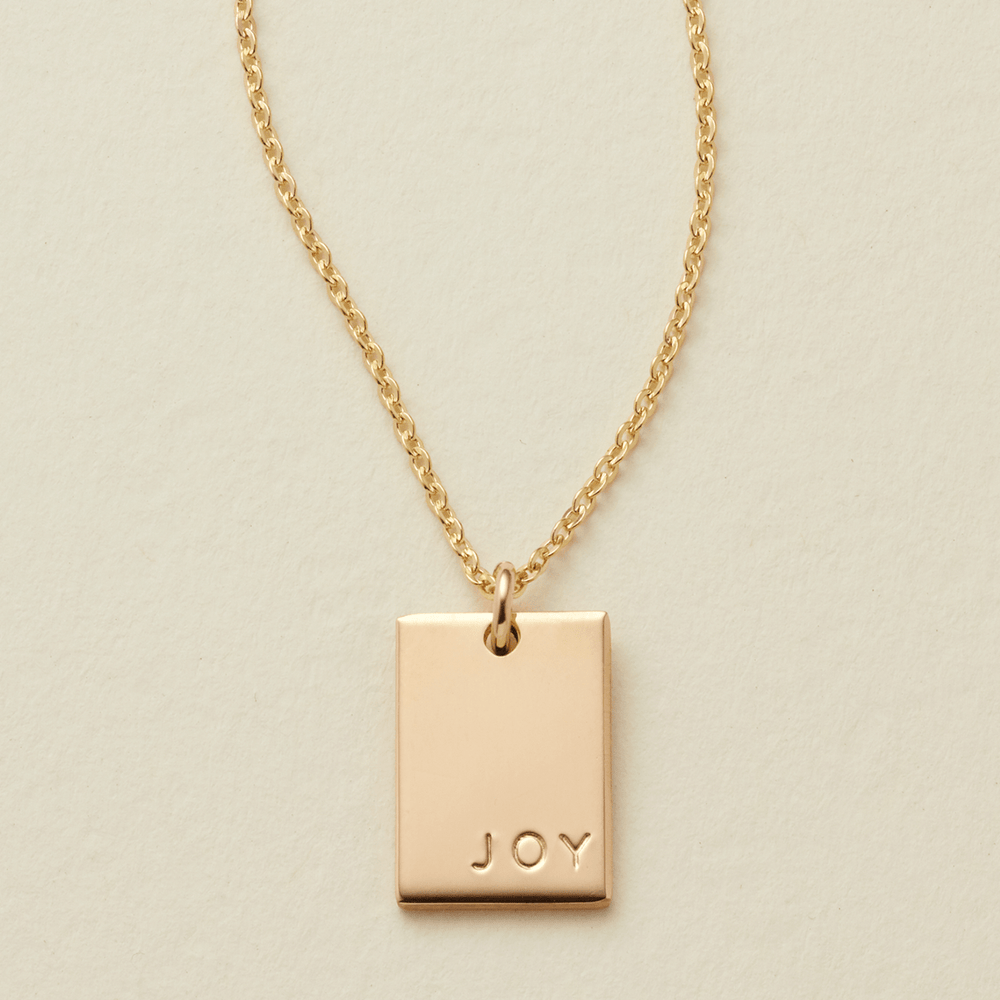 A gold chain necklace with a rectangular  gold pendant engraved with the word 'JOY', displayed on a textured light beige background.