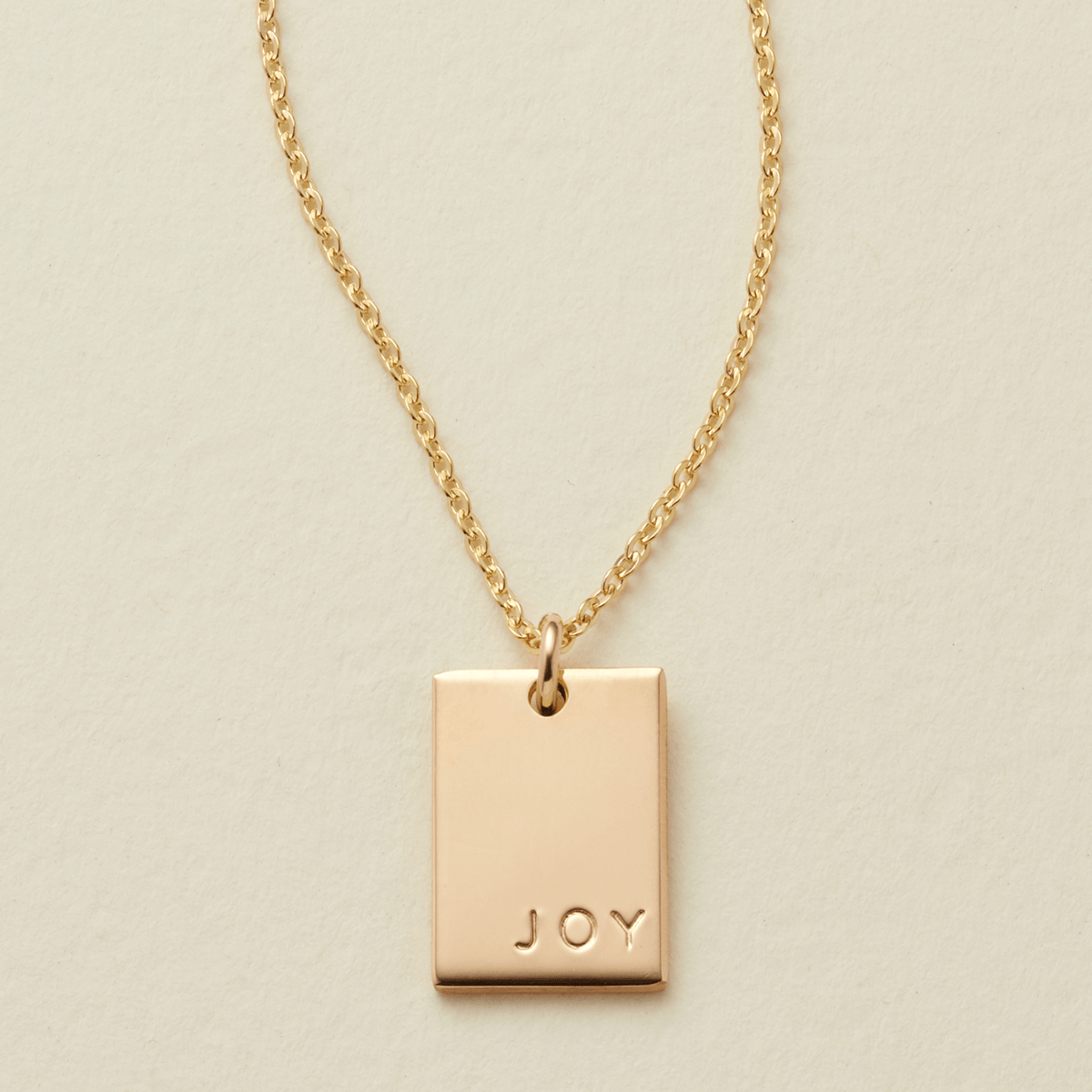 A gold chain necklace with a rectangular  gold pendant engraved with the word 'JOY', displayed on a textured light beige background.