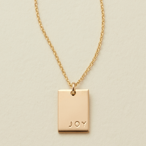 A gold chain necklace with a rectangular  gold pendant engraved with the word 'JOY', displayed on a textured light beige background.