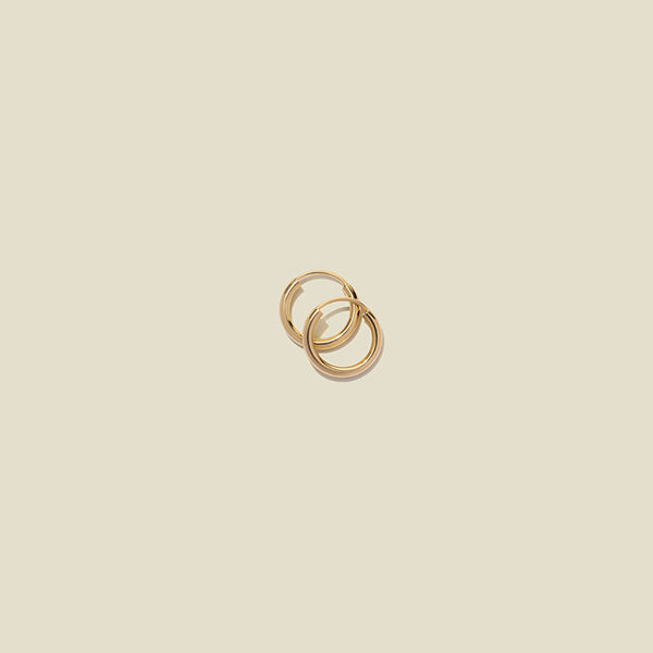 Two small thin gold hoop earrings laying on top of one another, shown on a light beige background.