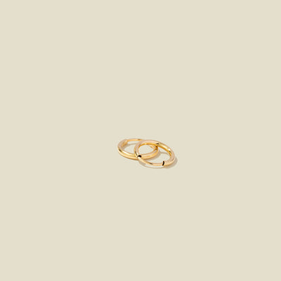 A pair of two small thin gold hoop earrings laying on top of one another, shown on a light beige background.