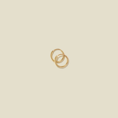 A pair of small thin gold hoop earrings laying on top of one another, shown against a solid light beige background.