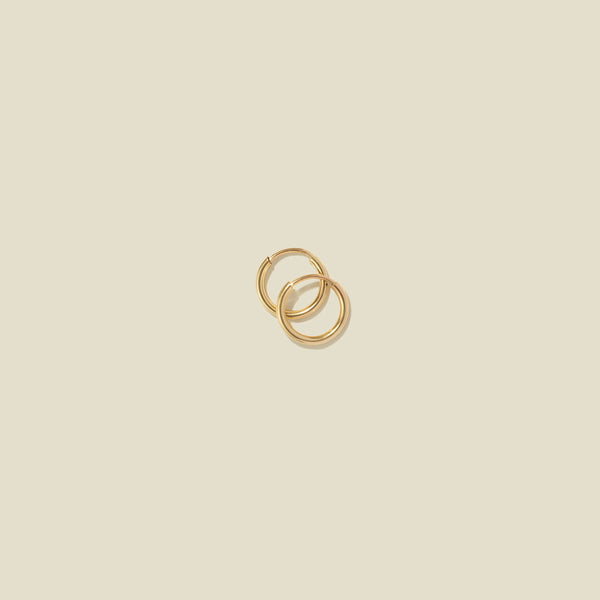 A pair of small thin gold hoop earrings laying on top of one another, shown against a solid light beige background.