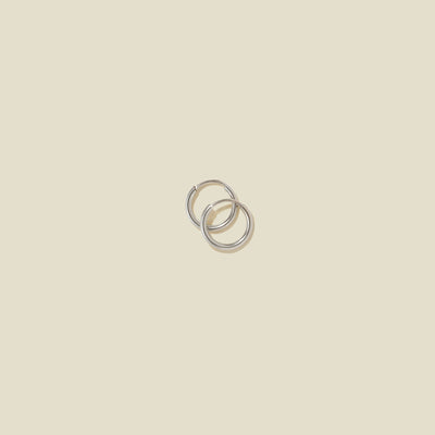 A pair of small thin silver hoop earrings laying on top of one another, shown on a solid light beige background.