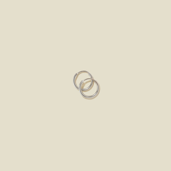 A pair of small thin silver hoop earrings laying on top of one another, shown on a solid light beige background.
