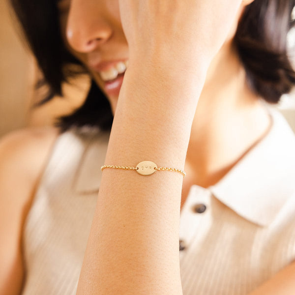 A close-up of a person's wrist wearing a gold chain bracelet with a flat oval set horizontally in the center, engraved with the letters 'CMR'.