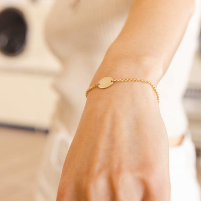 A close-up of a person's wrist wearing a gold chain bracelet with a flat oval set horizontally in the center, engraved with the letters 'CMR'.