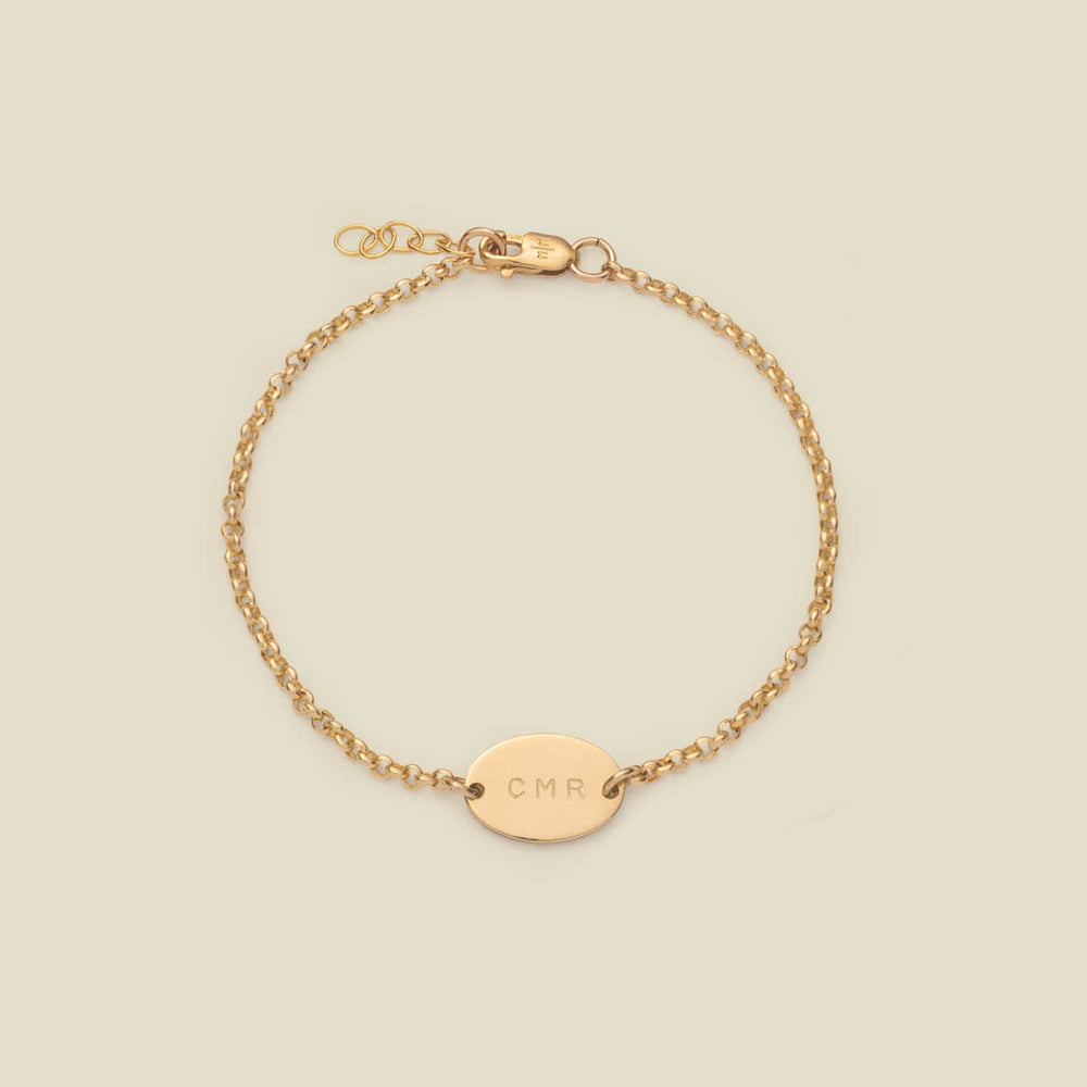 A gold chain bracelet with a flat oval charm horizontally in the center, engraved with the letters 'CMR', shown against a solid light beige background.