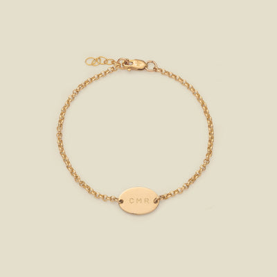 A gold chain bracelet with a flat oval charm horizontally in the center, engraved with the letters 'CMR', shown against a solid light beige background.
