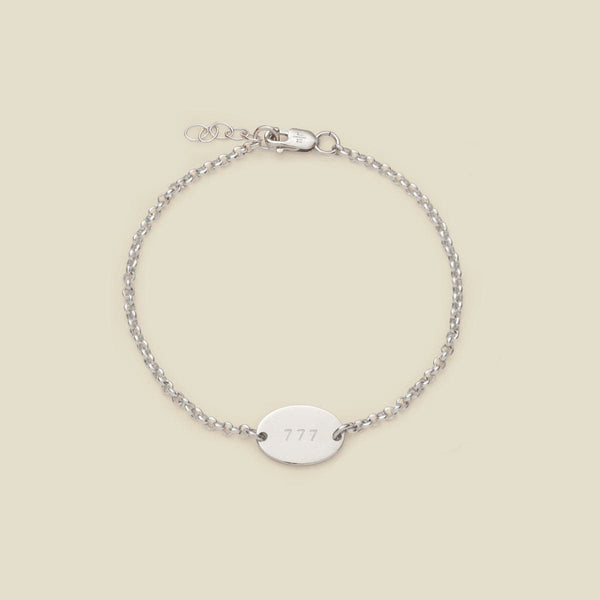 A silver chain bracelet with a flat oval set horizontally in the center, engraved with the letters '777', shown on a solid light beige background.