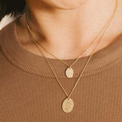 A close-up of a person in a brow ribbed top wearing two layered gold pendant necklaces, each with a different size of flat engraved oval disc.