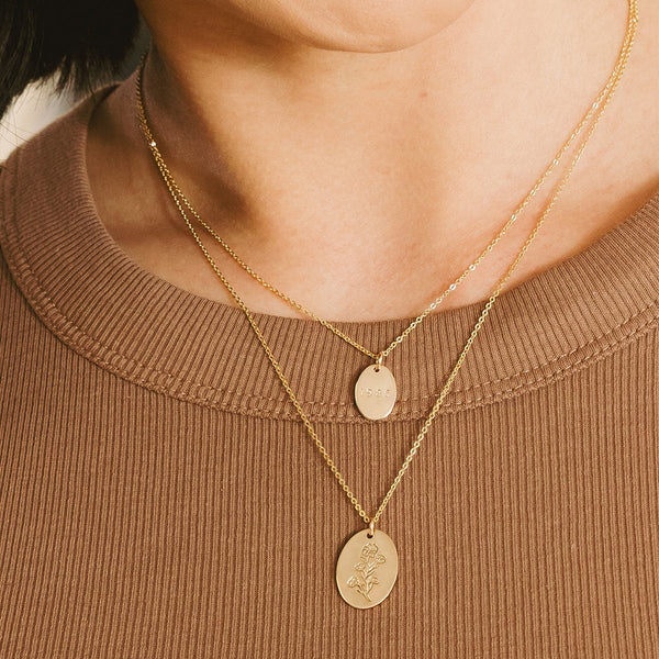A close-up of a person in a brow ribbed top wearing two layered gold pendant necklaces, each with a different size of flat engraved oval disc.