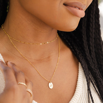 A close-up of a woman wearing a thin textured gold choker necklace layered with a long delicate pendant necklace featuring a small flat oval disc. The disc has a daisy floral motif engraved on it.