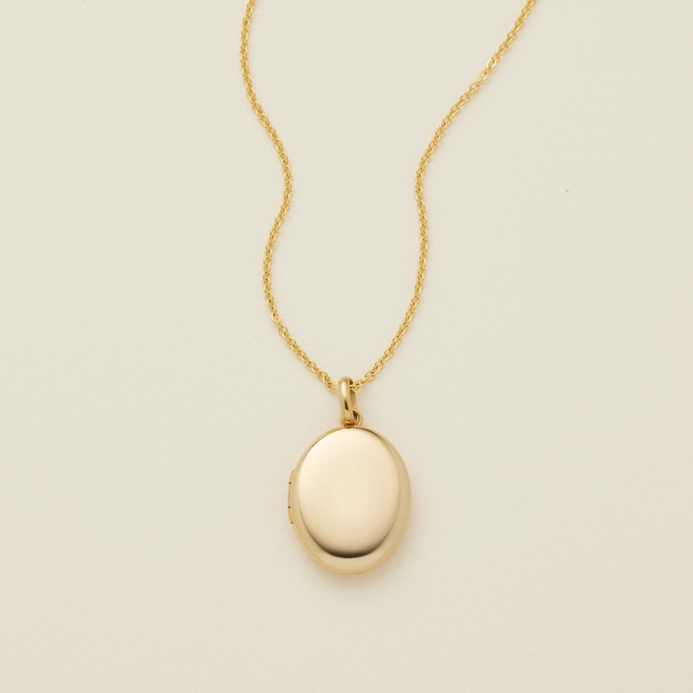 A gold oval locket necklace on a delicate chain, photographed against a solid off-white background.