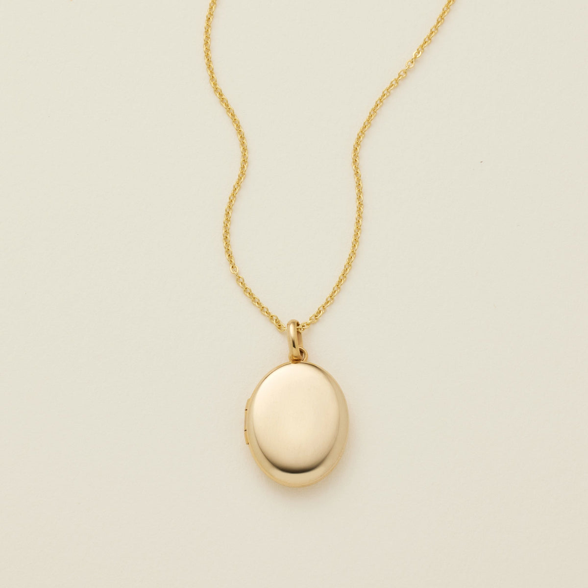 A gold oval locket necklace on a delicate chain, photographed against a solid off-white background.