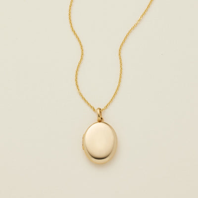 A gold oval locket necklace on a delicate chain, photographed against a solid off-white background.
