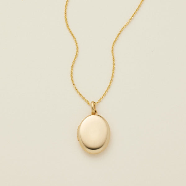 A gold oval locket necklace on a delicate chain, photographed against a solid off-white background.
