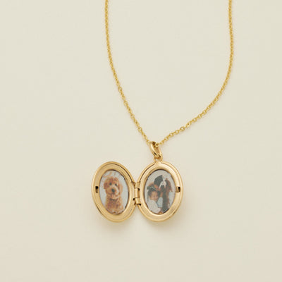 A gold oval locket necklace on a delicate chain, photographed open to show two photos inside, against a solid off-white background.