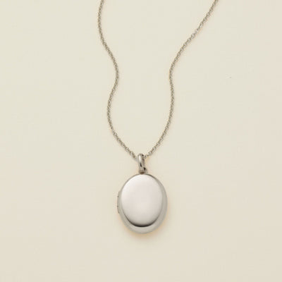 A silver oval locket necklace on a delicate chain, photographed against a solid off-white background.