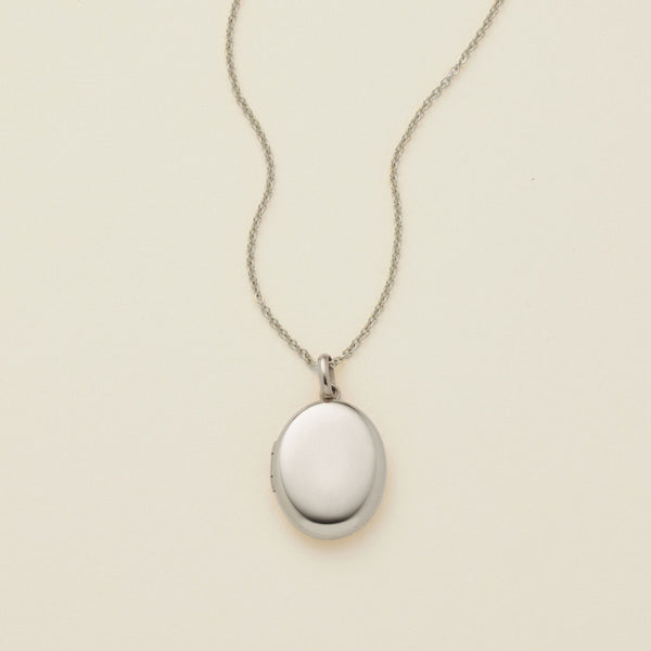 A silver oval locket necklace on a delicate chain, photographed against a solid off-white background.