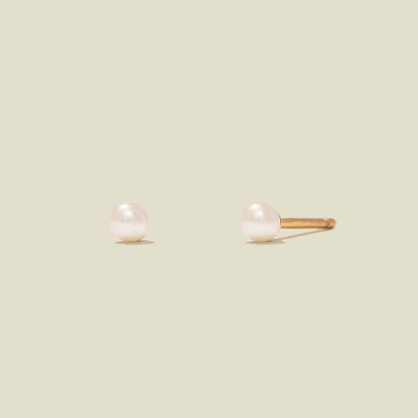 A pair of minimalist stud earrings featuring smooth, round white pearls on gold posts, set against a solid light beige background.