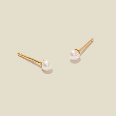 A pair of minimalistic gold studs with a small round white pearl, shown against a solid light beige background.
