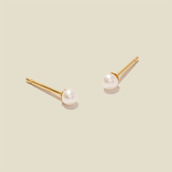 A pair of minimalistic gold studs with a small round white pearl, shown against a solid light beige background.