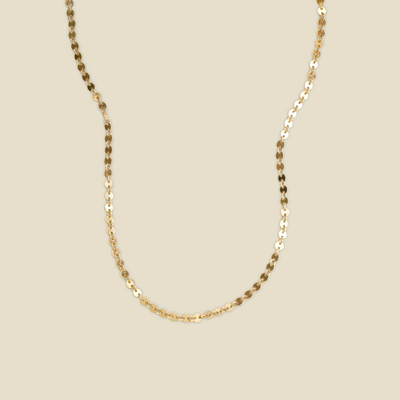 A delicate gold chain necklace with a textured, disc-like link pattern, shown draped against a solid beige background.