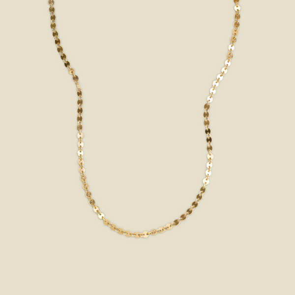 A delicate gold chain necklace with a textured, disc-like link pattern, shown draped against a solid beige background.