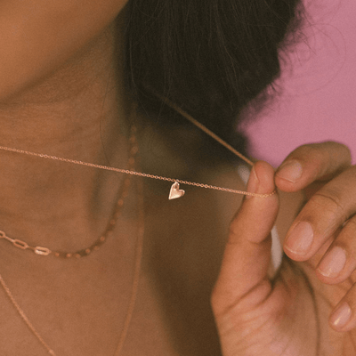 A person wearing multiple gold necklaces, holding a delicate pendant chain with a small flat gold heart charm, shown against a solid mauve background.