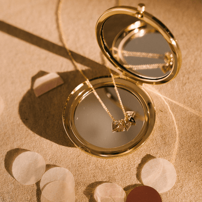 A gold compact mirror with a delicate gold pendant chain resting on the mirror. There are five flat gold heart charms on the chain, each engraved with a single initial. 