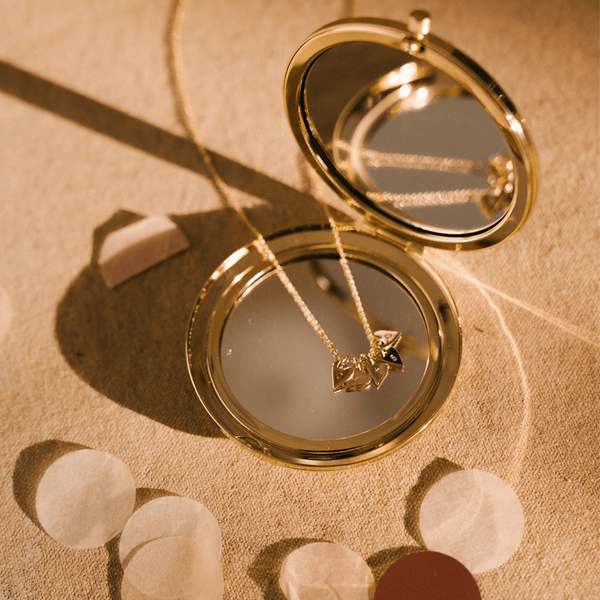 A gold compact mirror with a delicate gold pendant chain resting on the mirror. There are five flat gold heart charms on the chain, each engraved with a single initial. 