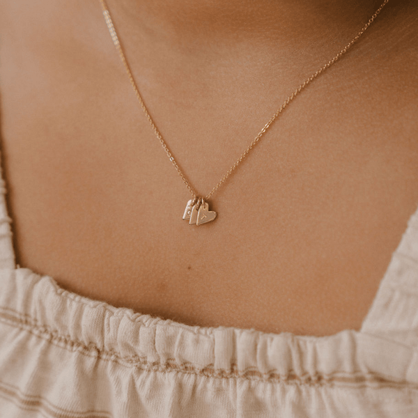 A close-up of a person wearing a delicate gold pendant chain with three small gold heart charms, each engraved with a single initial.