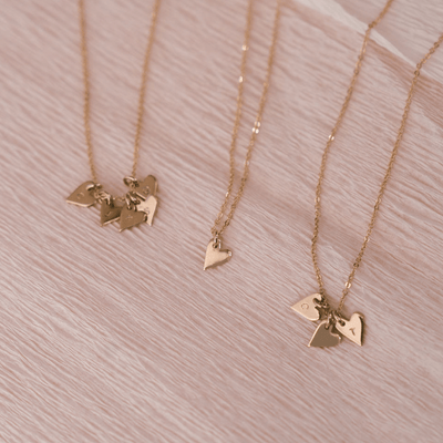 Three delicate gold pendant chain necklaces each with small flat heart charms. Some of the hearts are engraved with single initials, and all of the necklaces are laying on a textured light beige surface.